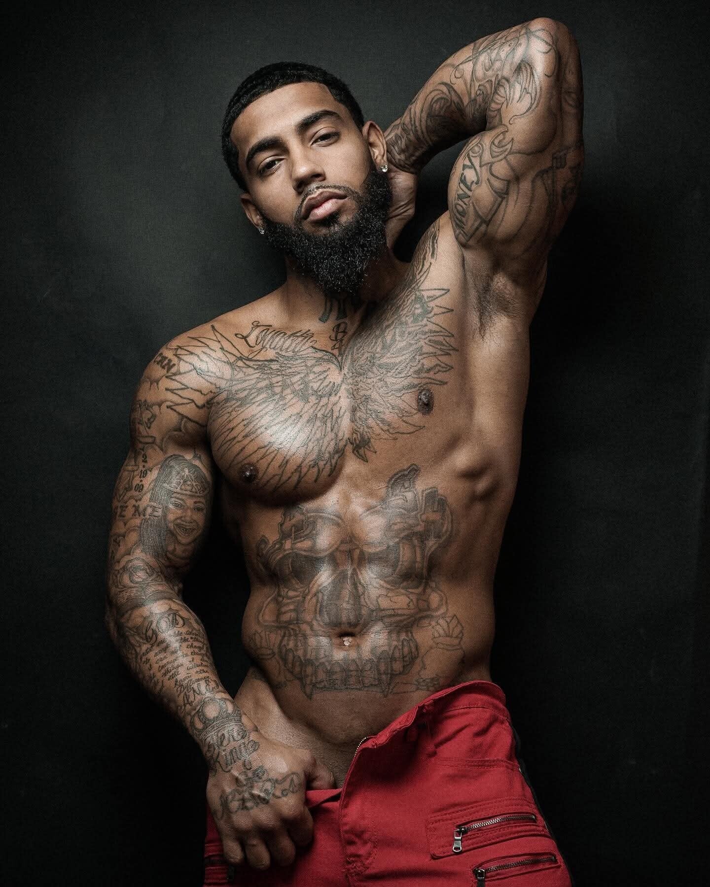 A handsome muscular, tattooed black & latin mixed man with a beard posing shirtless against a dark background, showcasing detailed chest and abdominal muscles, tattoos, and a confident alluring face expression.