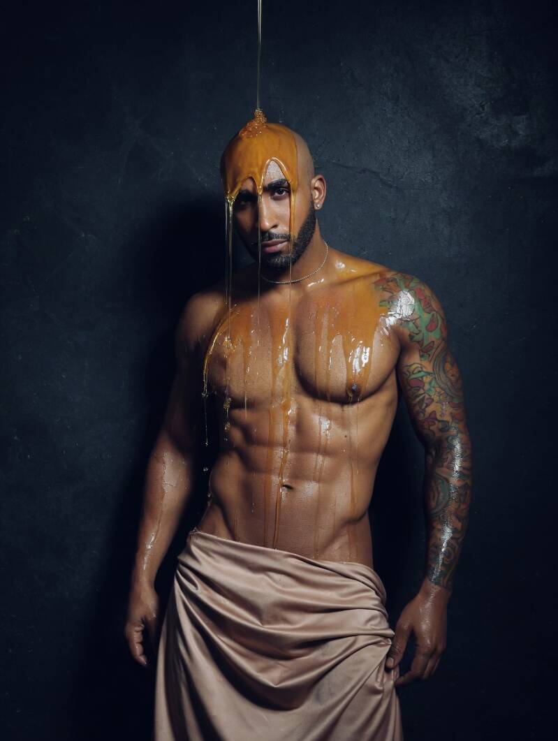 Artistic studio portrait of a fit, shirtless Black man wrapped in a tan cloth around his waist. He is looking down thoughtfully as a thick, golden substance pours over his head and down his chest.