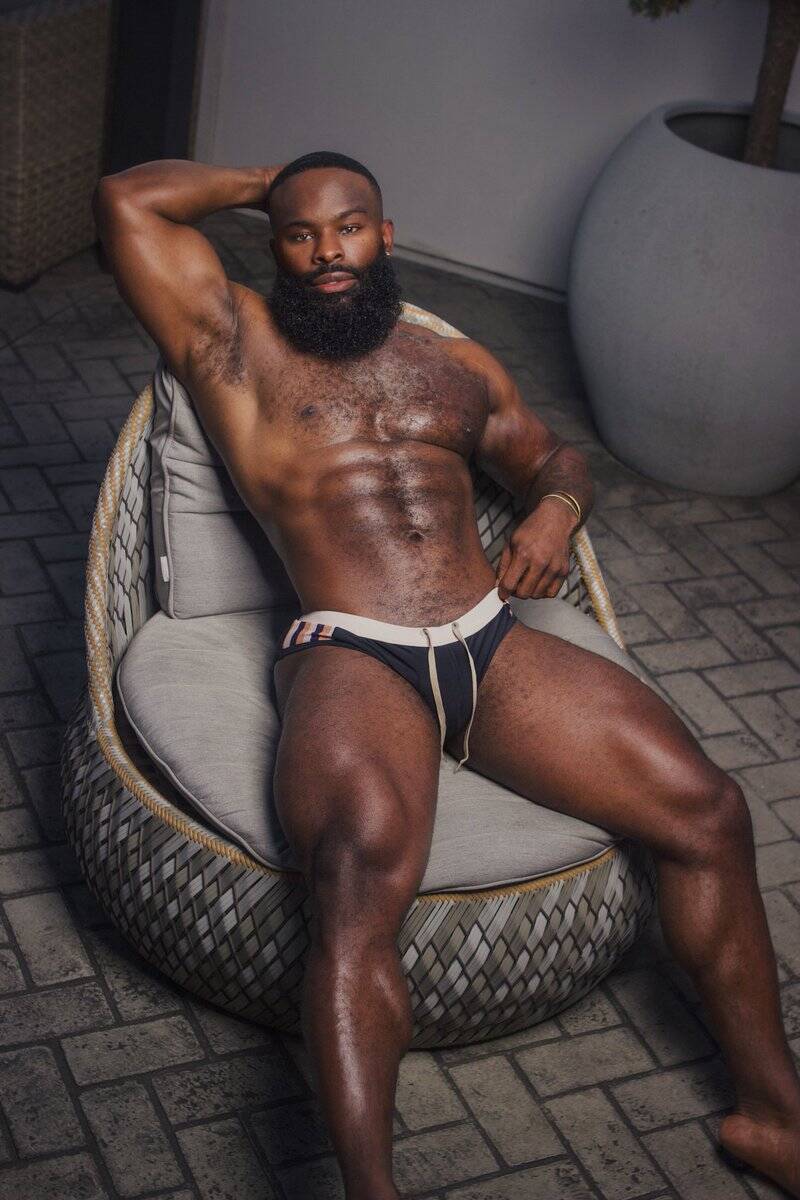 Sexy black male model lounging comfortably in a chair showing off defined shredded body, and toned legs in his underwear.