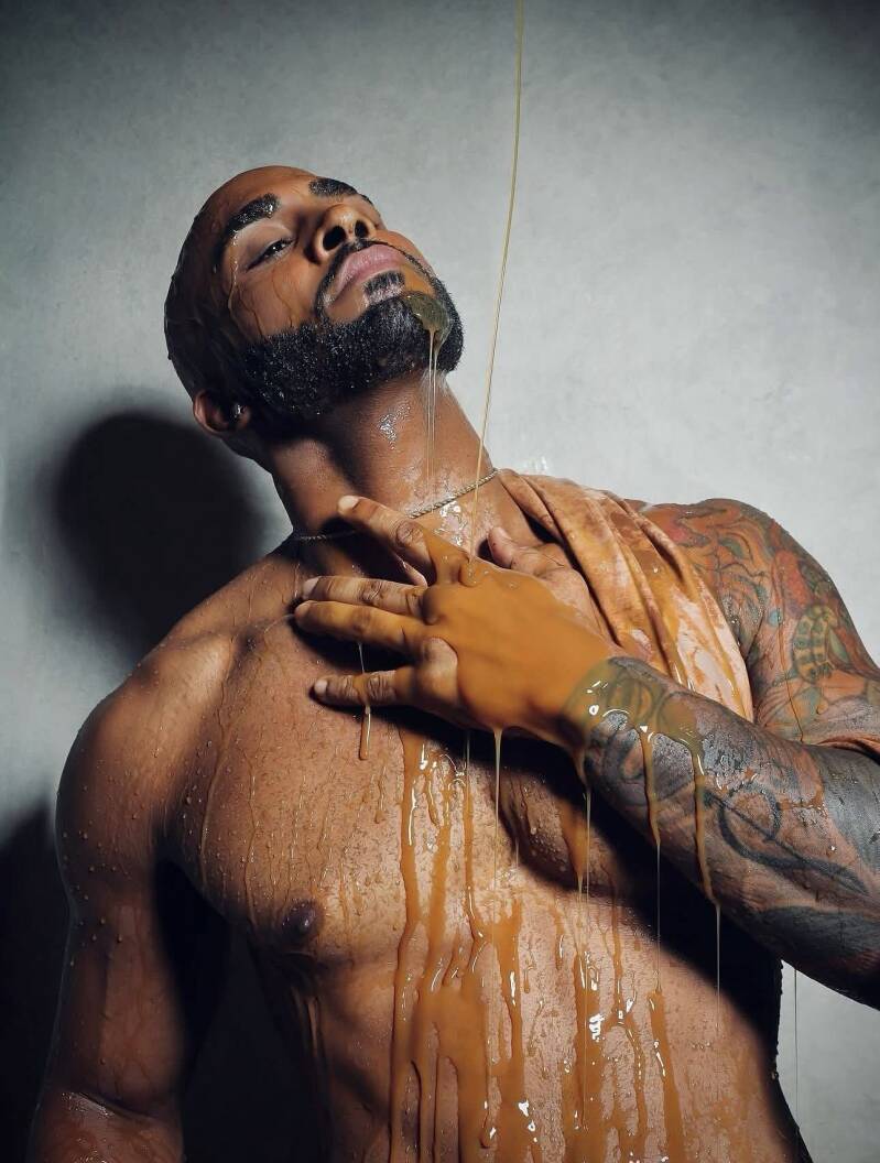 Low-angle close-up of a muscular Black man with head tilted back, using a cloth to wipe a viscous, golden liquid from his neck and tattooed arm in a dramatic studio lighting setup.