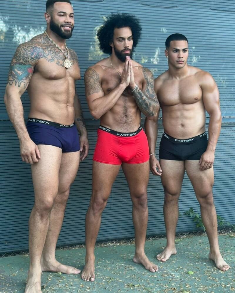 Three athletic, muscular, and barefoot Latino men with defined physiques pose outdoors showing off bodies and feet in blue, red, and black "FAST BOY" boxer briefs against a grey slatted wall.