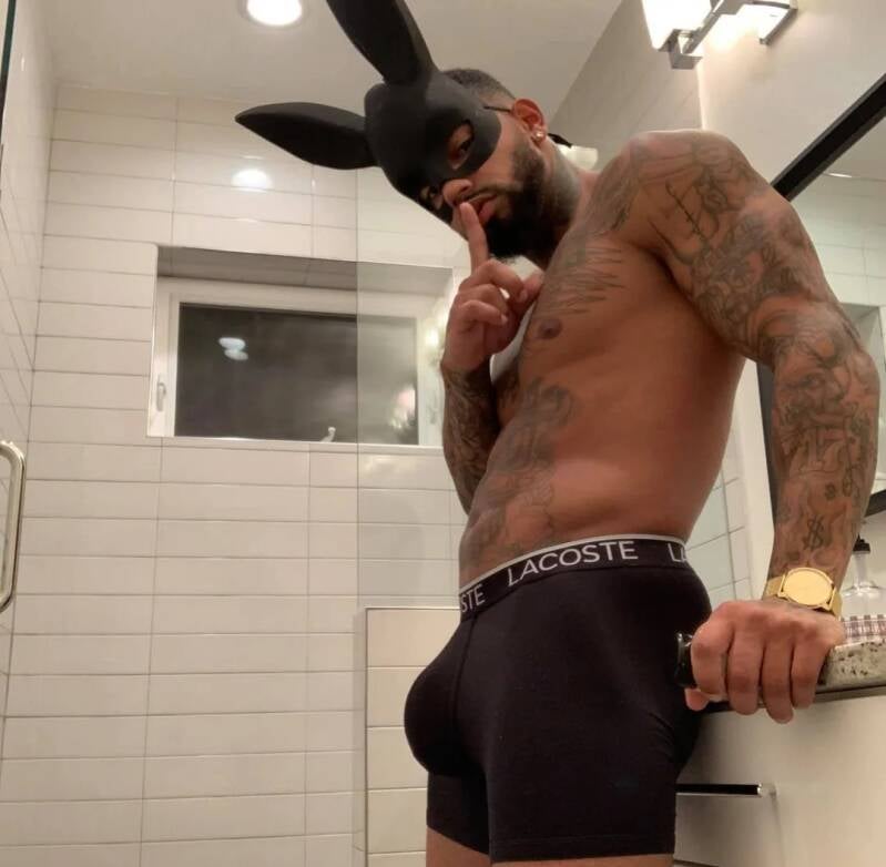 A muscular, tattooed man wearing a black bunny mask and black Lacoste boxer briefs poses in a bathroom, holding a finger to his lips. Looking alluring and sexy. He looks as though he's about to role play.
