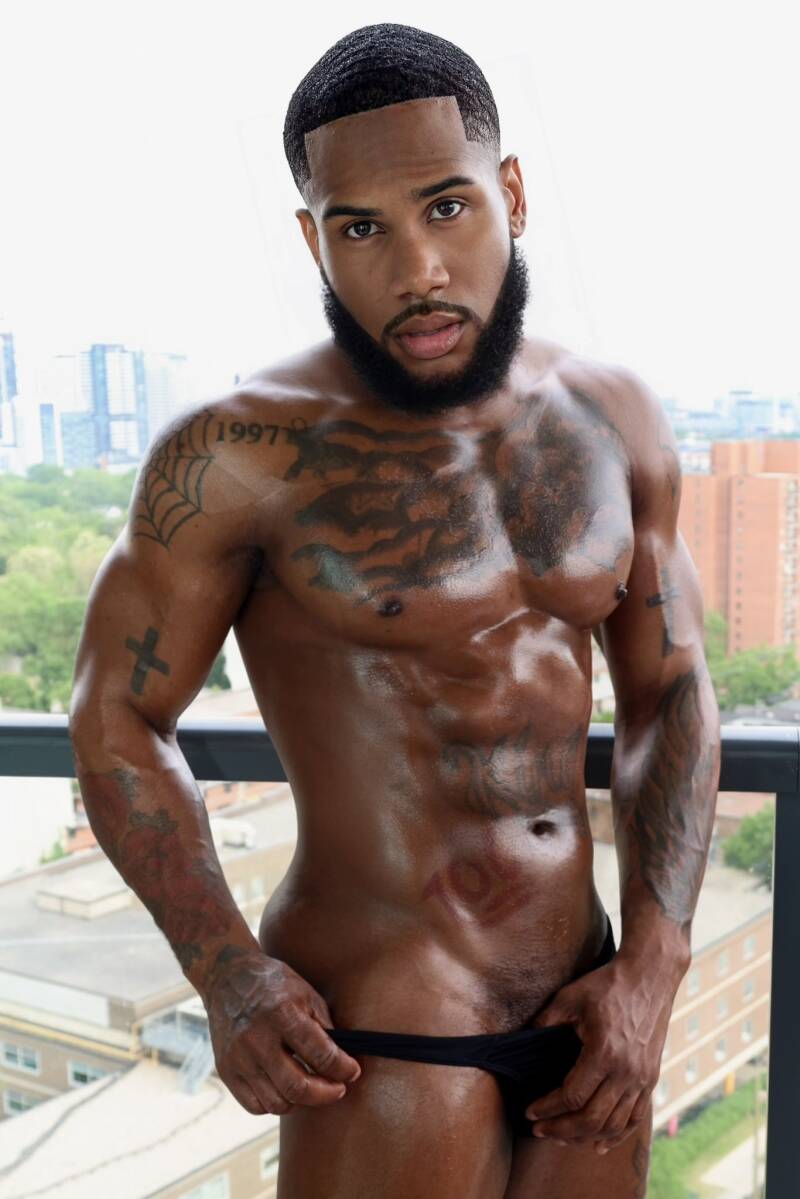 A hot, muscular Black man with short hair, a full beard, and chest tattoos posing shirtless  modeling his hot physique on an urban balcony with a city background.