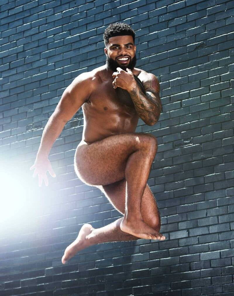 Ezekiel A hot, muscular Black athlete with a handsome smile and full beard jumping mid-air. He showcases a shredded athletic body type, defined abs, and bare feet while holding a football against a dark brick wall backdrop.
