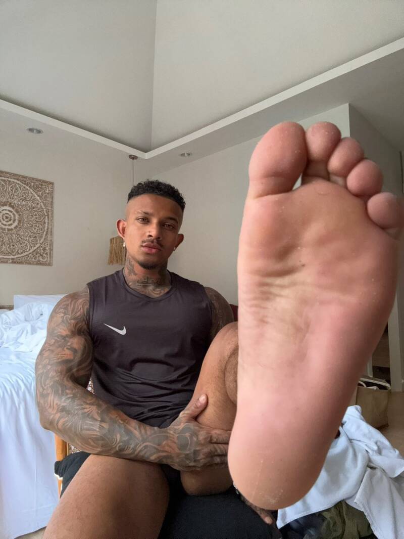 Close-up indoor view of a tattooed sexy Latin man's right sole. The sole shows a rugged, natural texture with mild dry patches and slight skin flaking. These are especially visible around the outer edges of the heel, and the base of the toes.