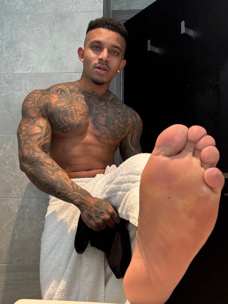 Close-up foreground view of a tattooed muscular Latin man's smooth right sole, showing thick rounded toes and a wide athletic foot structure while wrapped in a white towel.