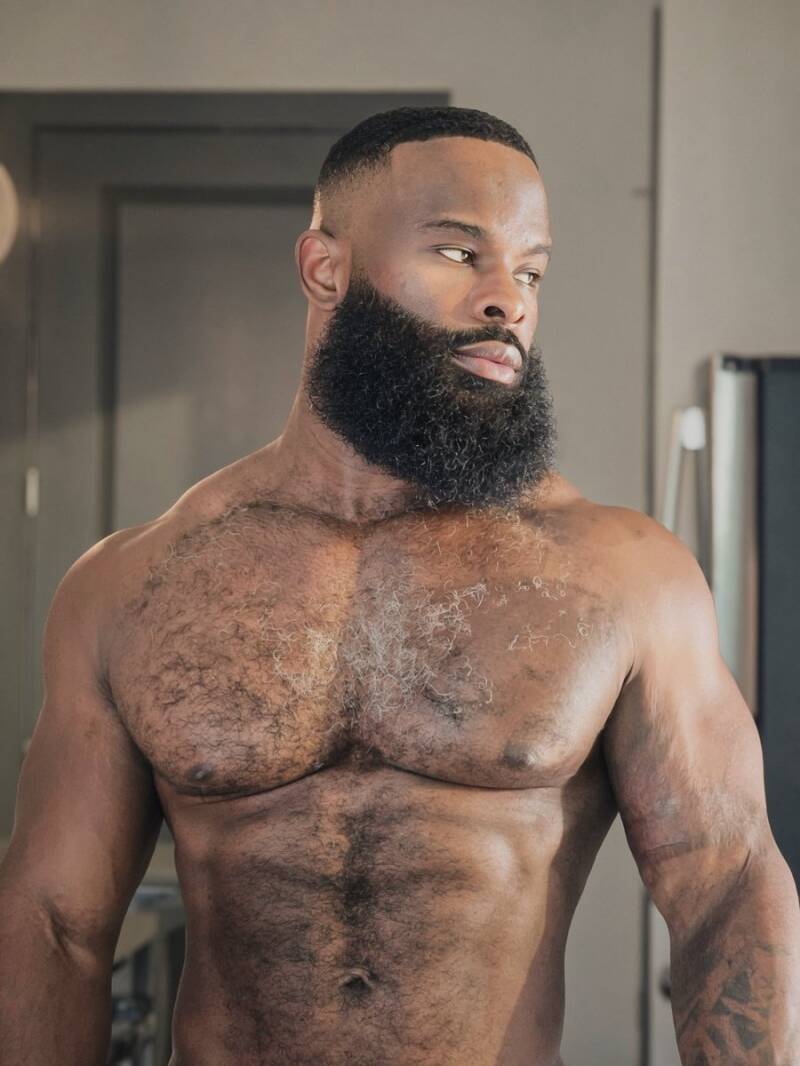 Up-close image of a tall, dark, and handsome shirtless black male model with thick groomed beard, and chiseled muscular physique with chest and stomach hair, making him look confident, masculine and strong.