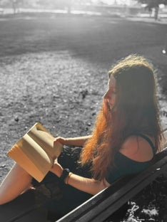 Das Bild zeigt mich seitlich im Profil auf einer Bank sitzend. Ich habe ein aufgeschlagenes Buch in den Händen und lese darin. Im Hintergrund ist eine ausgegraute Wiese.