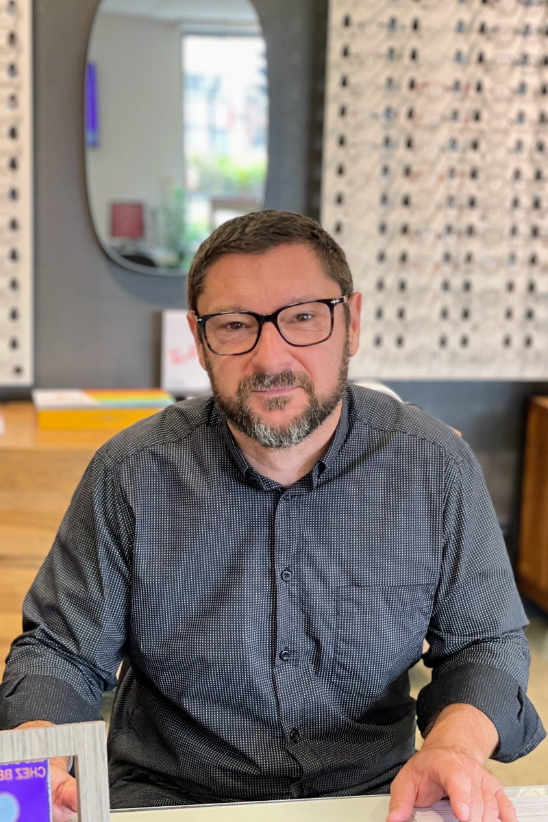 Fabien Roussel, Employé de la boutique Moderne Optique assis à un bureau, portant des lunettes et une chemise à motifs discrets. En arrière-plan, un grand miroir ovale, des étagères de lunettes exposées, et une fenêtre laissant entrer la lumière naturelle