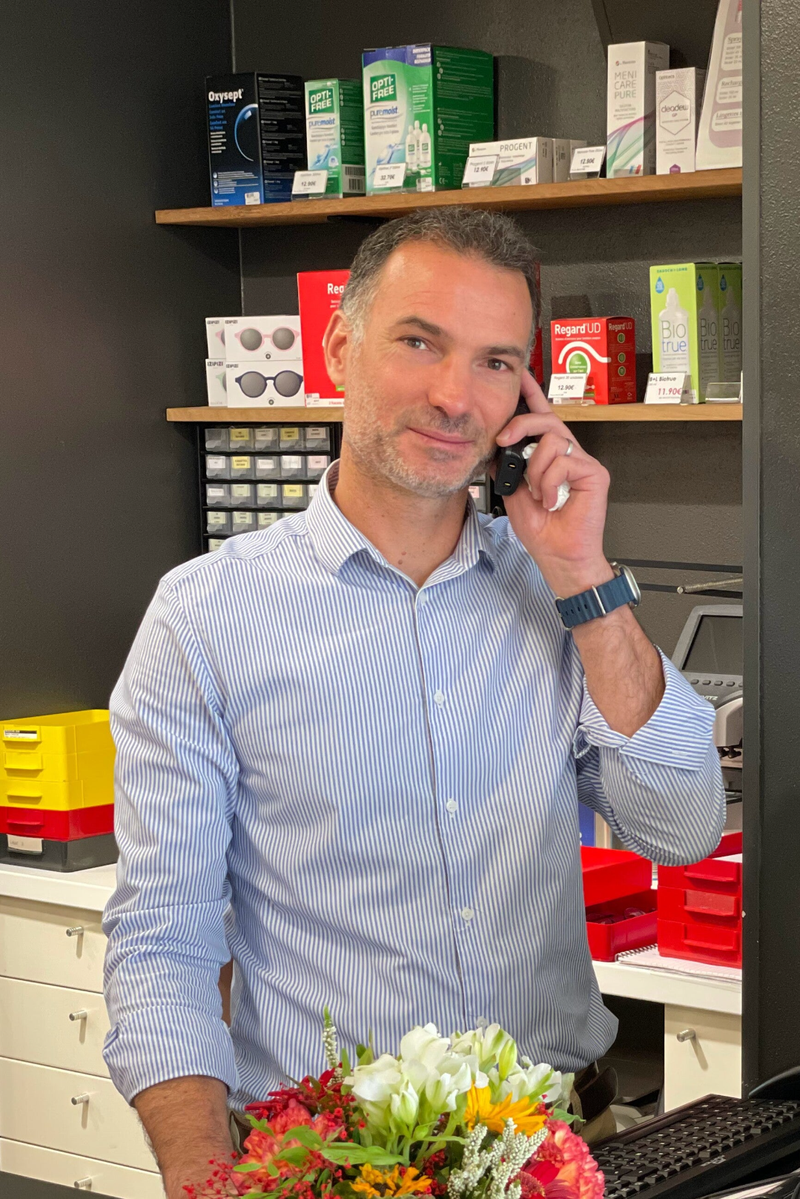 Lionel Grande fondateur de la boutique Moderne Optique en train de téléphoner derrière le comptoir, entouré de produits d'optique et de soins pour lentilles disposés sur des étagères. Un bouquet de fleurs colorées est posé devant lui, ajoutant une touche