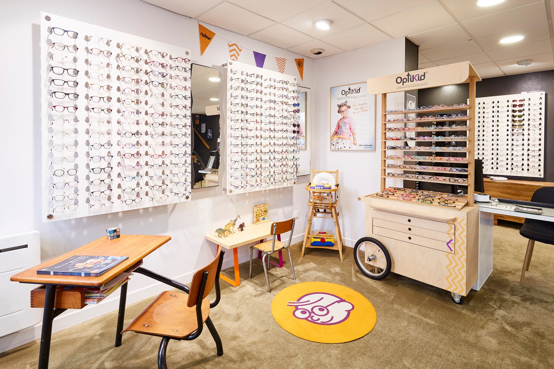 Affiche OptiKid, spécialiste de la vue des enfants, montrant un jeune garçon portant des lunettes et une petite fille en bas âge avec des lunettes roses. Le slogan 'Nos lunettes, une affaire de spécialiste !' est inscrit en orange à côté des enfants. En b