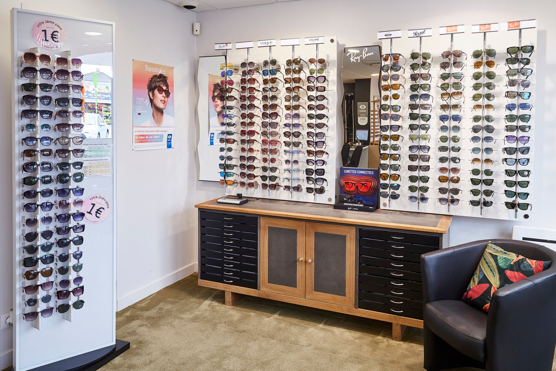 Intérieur de la boutique Moderne Optique avec un présentoir de lunettes Ray-Ban exposant une large gamme de montures. À côté, un meuble à tiroirs noirs et des affiches de test de vue accrochées au mur. Une table avec un écran et des chaises noires est pla