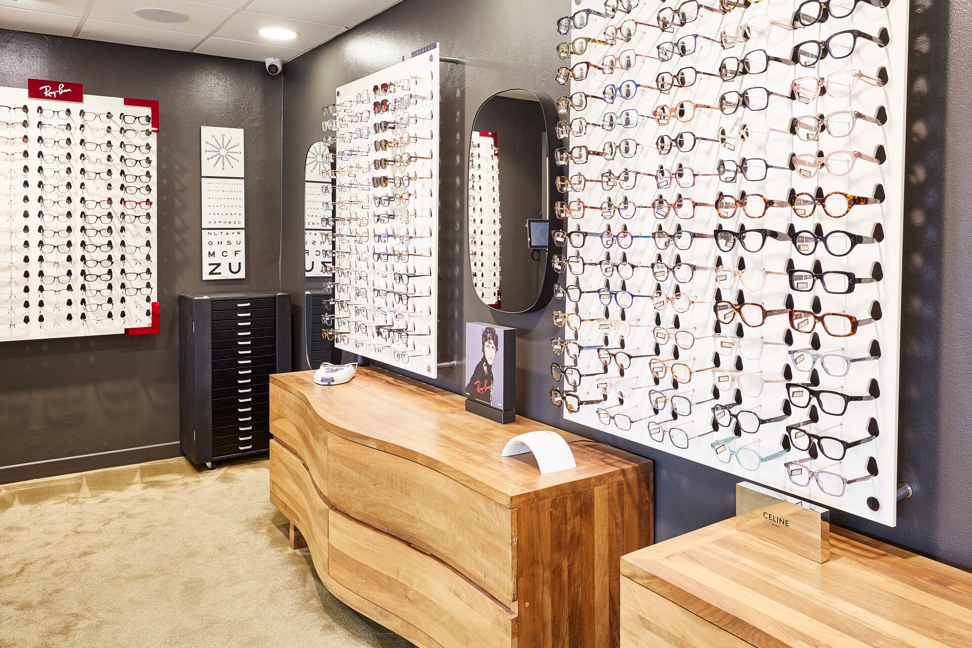Intérieur de la boutique Moderne Optique montrant un présentoir de lunettes de soleil avec des montures de différentes marques, y compris Ray-Ban, disposées sur un mur. Un meuble bas en bois avec des tiroirs noirs se trouve sous le présentoir, et deux fau