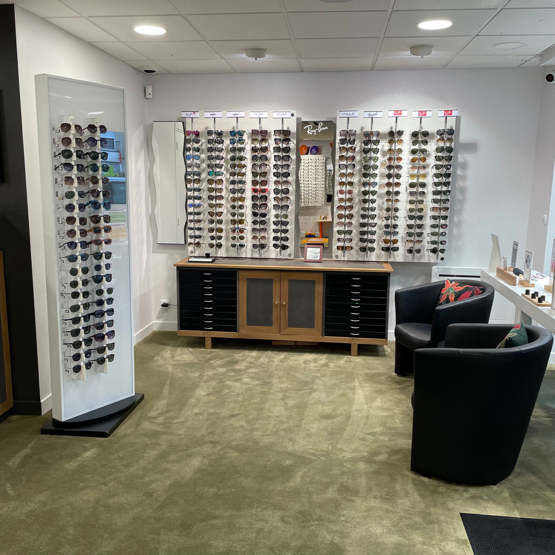 Intérieur de la boutique Moderne Optique montrant un présentoir de lunettes de soleil avec des montures de différentes marques, y compris Ray-Ban, disposées sur un mur. Un meuble bas en bois avec des tiroirs noirs se trouve sous le présentoir, et deux fau