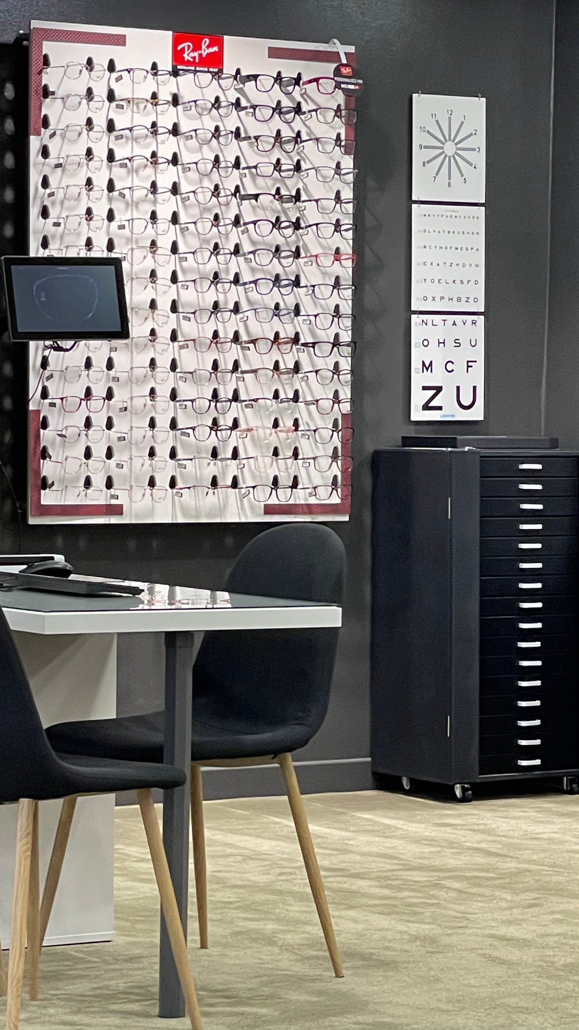 Intérieur de la boutique Moderne Optique avec un présentoir de lunettes Ray-Ban exposant une large gamme de montures. À côté, un meuble à tiroirs noirs et des affiches de test de vue accrochées au mur. Une table avec un écran et des chaises noires est pla