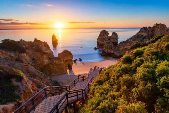 Wooden stairs descending to Praia do Camilo in Lagos, Portugal, with golden cliffs, offshore rock formations and warm sunlight over the sea.