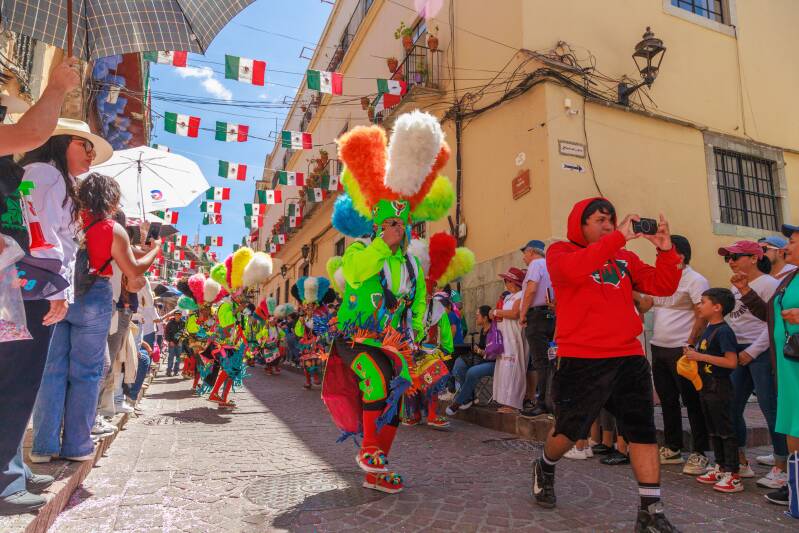 Street parade in Mexico with dancers wearing colourful feathered headdresses, spectators and Mexican flags strung above a cobbled street.