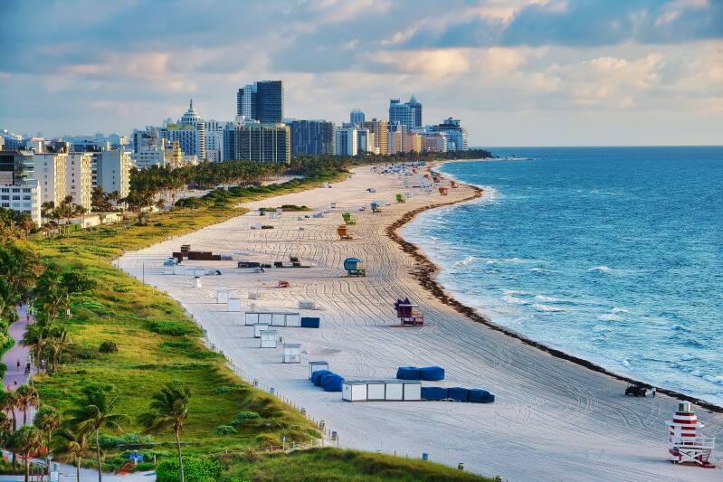 Wide sandy beach in Florida with colourful lifeguard towers, palm trees, ocean waves and beachfront high-rise buildings.