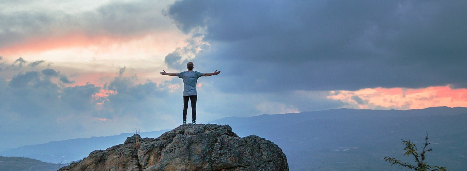op de top van een berg staat iemand met uitstrekte armen de beleving het uitzicht in zich op te nemen