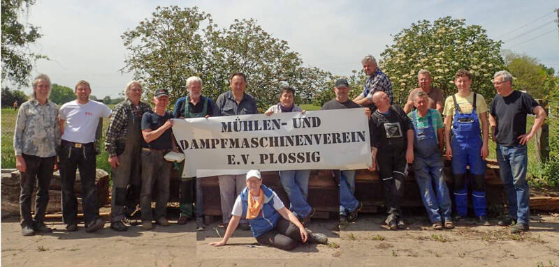 Vereinsmitglieder des Mühlen- und Dampfmaschinenvereins posieren für ein Foto auf dem Mühlenhof. Einige Personen halten ein Banner mit dem Vereinsnamen hoch. Im Hintergrund befinden sich Büsche und Bäume.