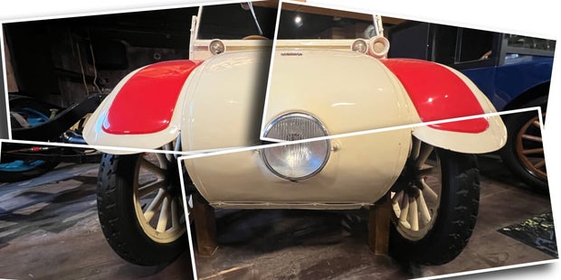 Frontansicht eines historischen Fahrzeugs mit cremefarbenem Karosseriekörper, roten Kotflügeln und einem zentralen runden Scheinwerfer, ausgestellt in einem Museum.