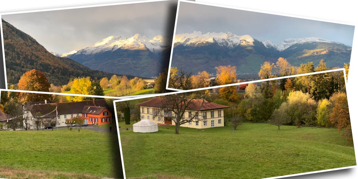 Malerische Hügellandschaft mit herbstlich gefärbten Bäumen, KLostergebäuden im Vordergrund und schneebedeckten Alpen im Hintergrund unter bewölktem Himmel.
