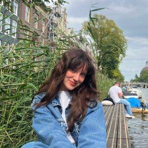 Een jongvolwassen vrouw met lang bruin haar zit buiten op een bank en lglimlacht voor zich uit.