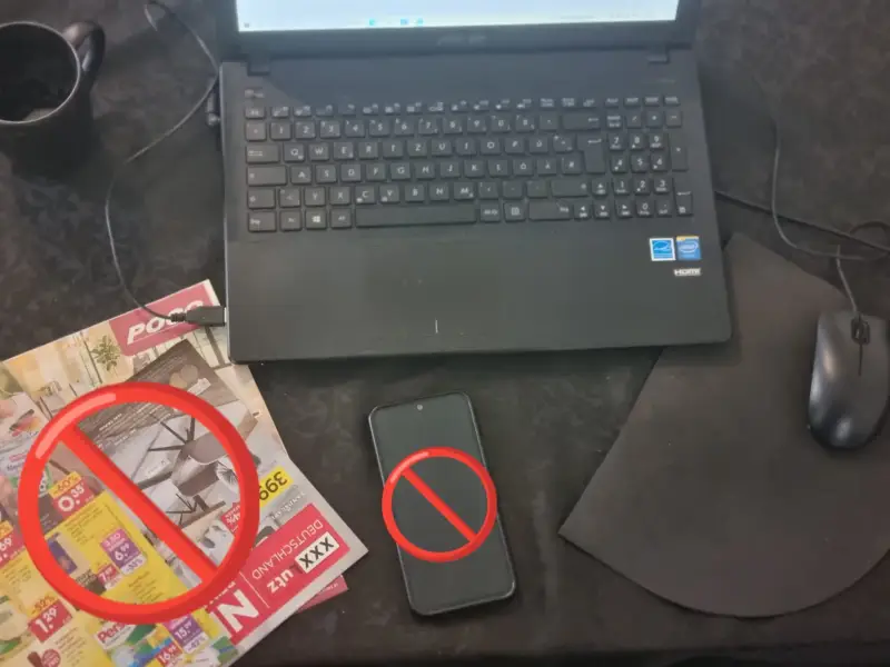 Ein Schreibtisch im Homeoffice mit einem Laptop, einem Smartphone und bunten Werbeprospekten, die als typische Ablenkung die Produktivität stören.