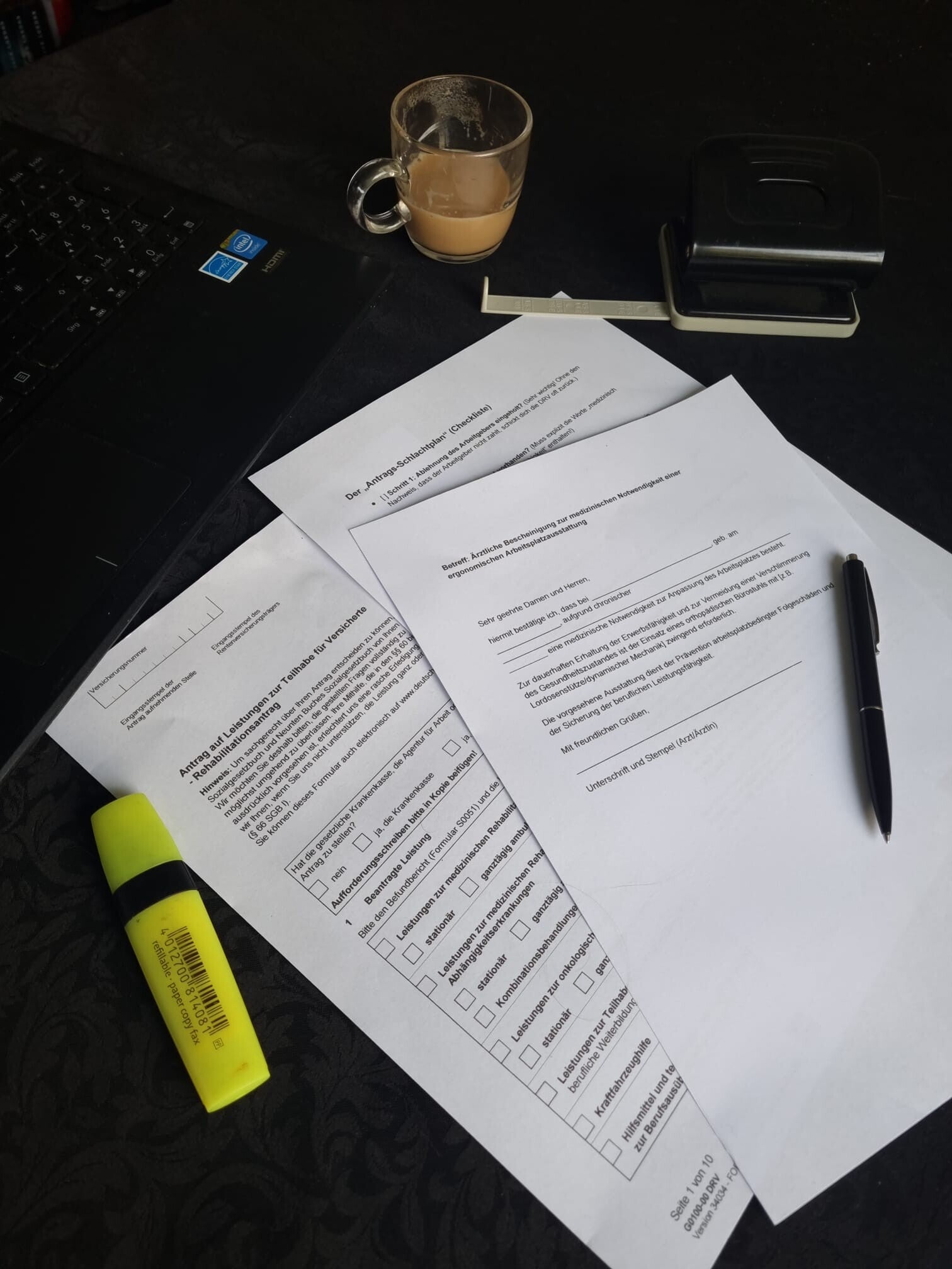Physische Antragsunterlagen der Deutschen Rentenversicherung und Krankenkasse für einen orthopädischen Bürostuhl auf einem Schreibtisch mit Stiften und Locher.