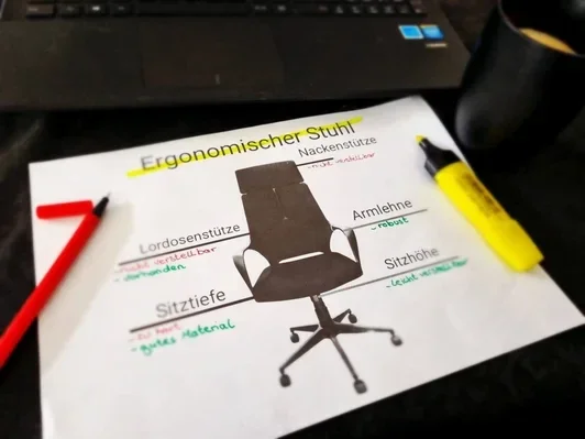 Detaillierte Analyse eines ergonomischen Bürostuhls auf gedrucktem Papier mit handgeschriebenen Anmerkungen und Checklisten-✓-Zeichen für Nackenstütze, Lordosenstütze, Sitzteife, Armlehne und Sitzhöhe. Im Hintergrund unscharf ein Laptop und einen Kaffee.