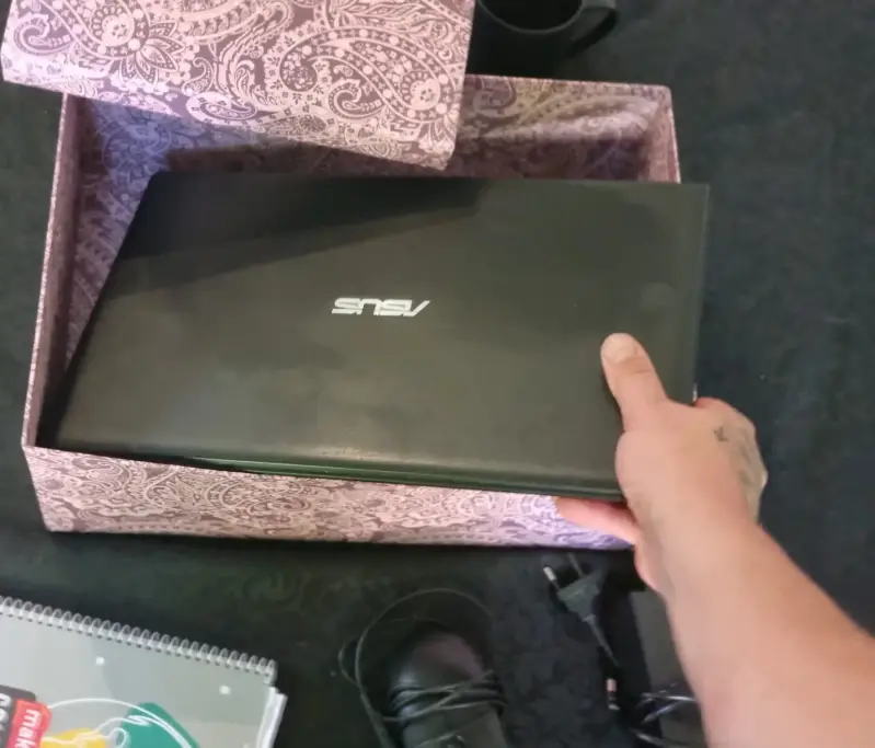 Ein schwarzer Laptop wird nach Feierabend zusammen mit einer Maus in eine gemusterte Bastelbox gelegt, um das Büro-Chaos im Wohnzimmer schnell zu verstauen.