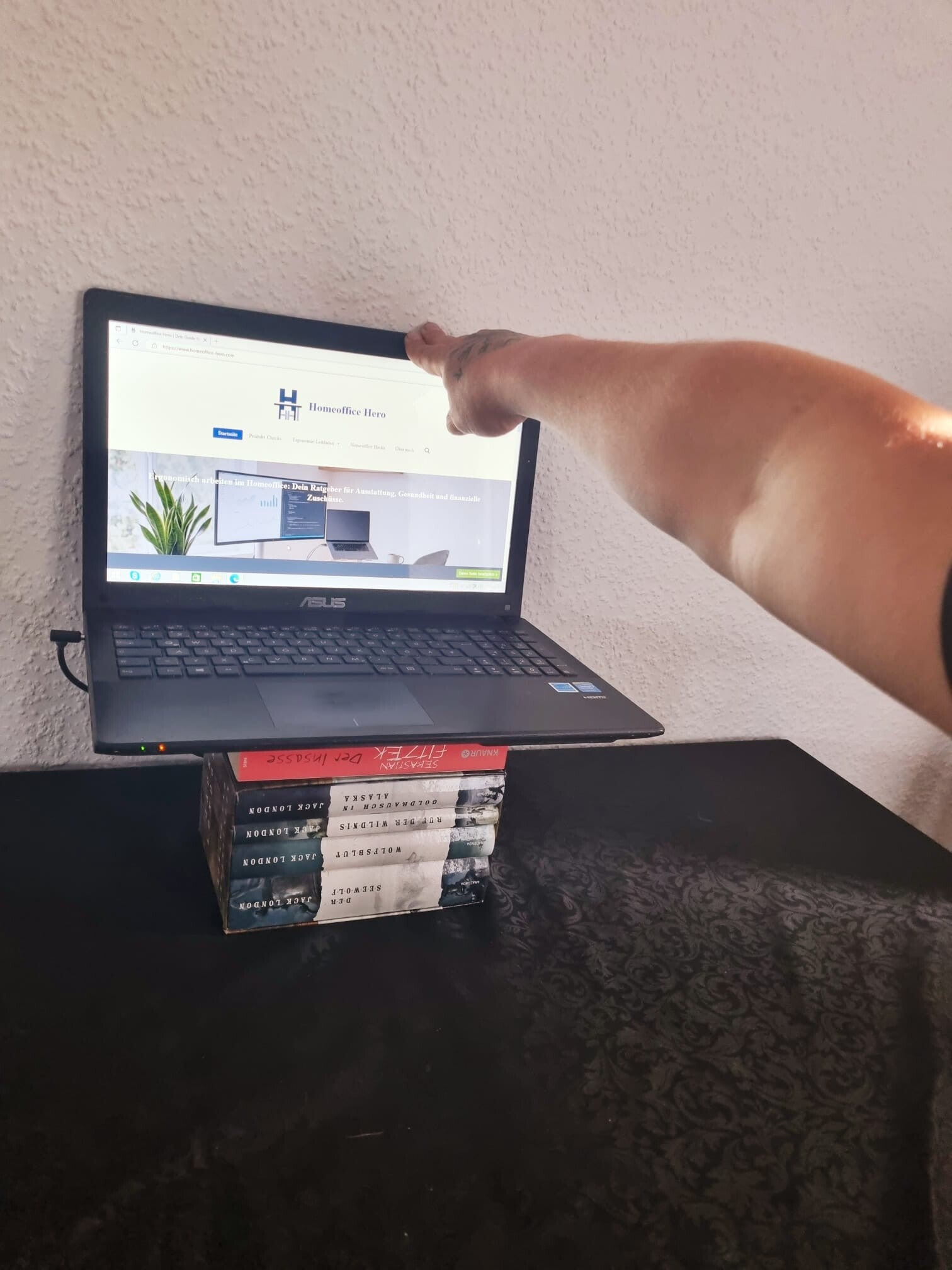 Ein Arm der an den oberen drittel eines Laptops zeigt und somit die Entfernung von Bildschirm zu Gesicht optimal einstellt. Der Laptop steht auf Büchern.