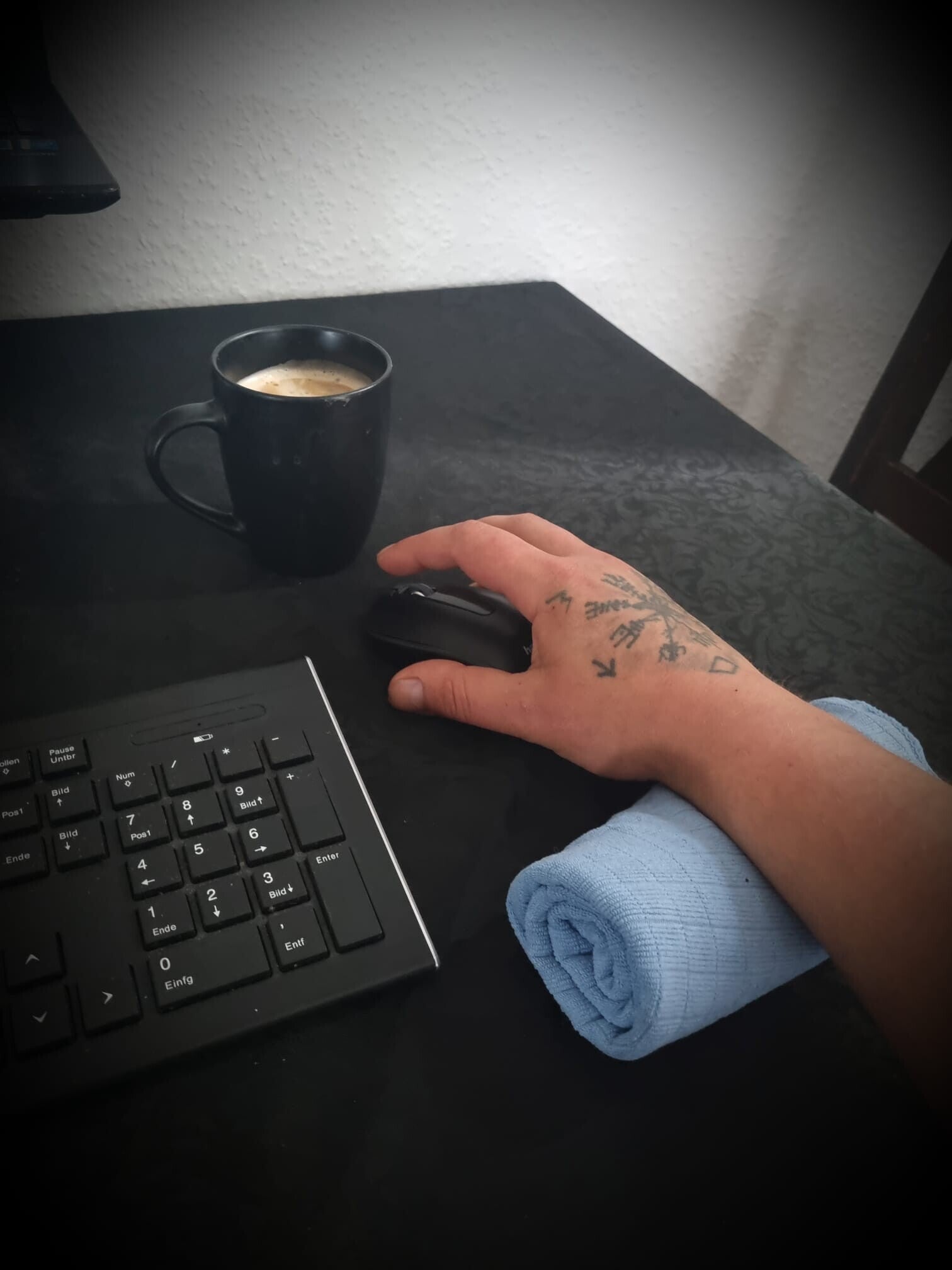 Eine tättowierte Hand auf einer Maus und zur entlastung ein gefaltetes Handtuch unter dem Handballen. Daneben eine Tastatur und eine Kaffeetasse mit Kaffee.