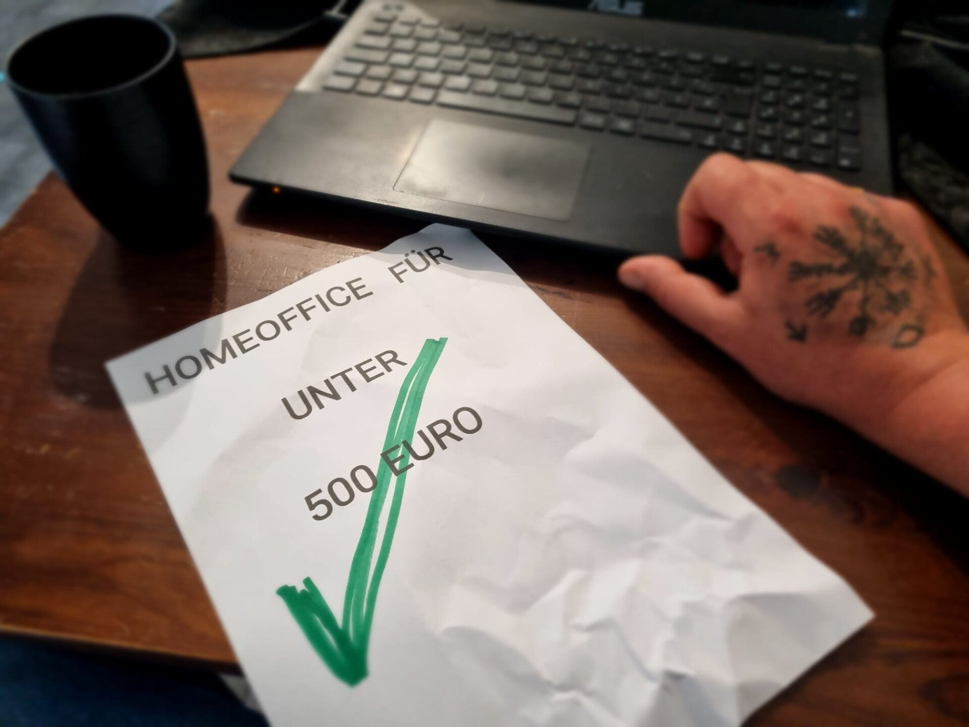 Eine tättowierte Hand, eine leere Kaffeetasse, ein Laptop und ein Zettel wo drauf steht:Homeoffice für unter 500 Euro und mit einem grünen Haken abgehakt.