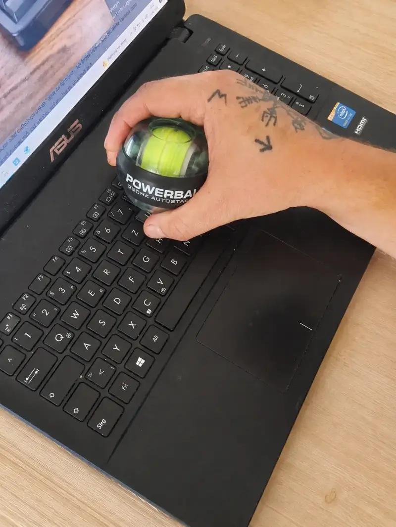 Eine Hand hält einen rotierenden Powerball (Handgelenktrainer) über der Tastatur eines Asus-Laptops. Das Gadget wird für eine kurze, aktive Unterarm-Übung direkt am Schreibtisch im Homeoffice genutzt, um Mausarm-Beschwerden vorzubeugen.