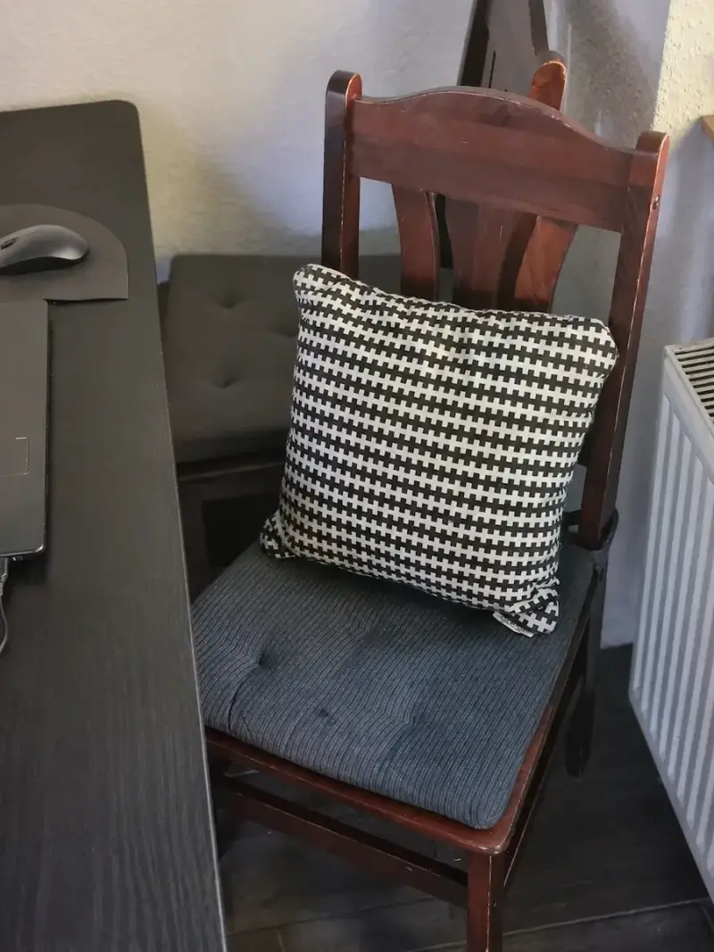 Ein einfaches Sofakissen auf einem Holzstuhl am Schreibtisch, das im Homeoffice als kurzzeitiger Ersatz für ein echtes Lordosenstütze Kissen genutzt wird.
