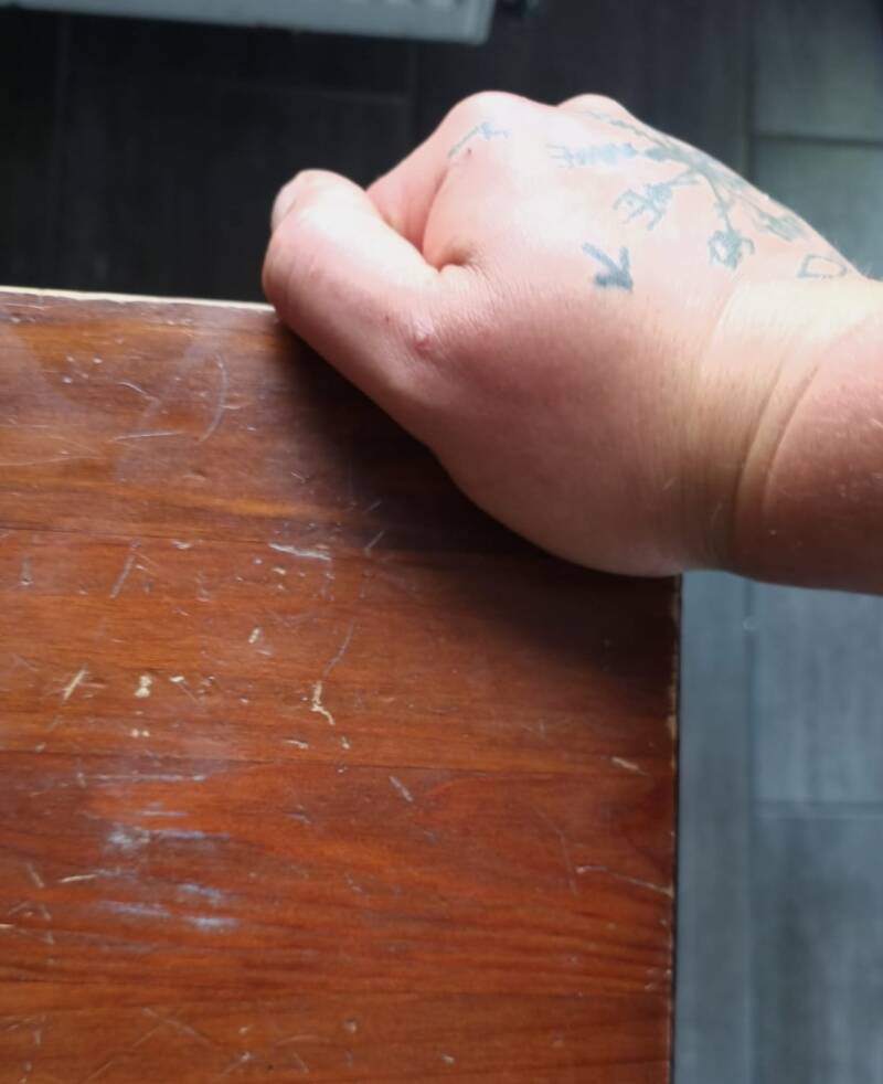 Eine Hand die auf einer Ecke eines Schreibtisches wackelt, um die Stabilität zu testen. Die Hand ist tättowiert und man sieht noch einen Teil des Bodens.