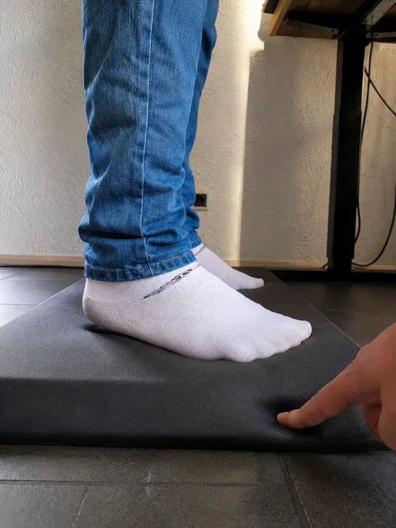 Praxistest einer Anti-Ermüdungsmatte im Homeoffice: Ein Finger drückt in das weiche Material, um die Dämpfung zu demonstrieren, während jemand in Socken am Stehschreibtisch steht.