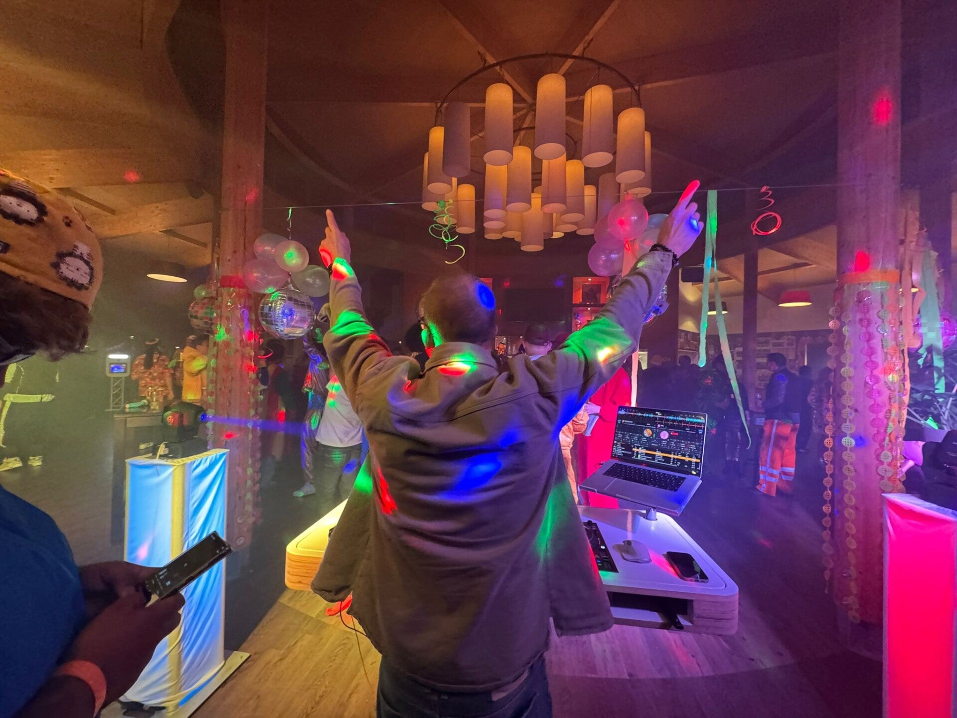 DJ Peter met zijn handen in de lucht achter de draaitafel, omringd door feestverlichting