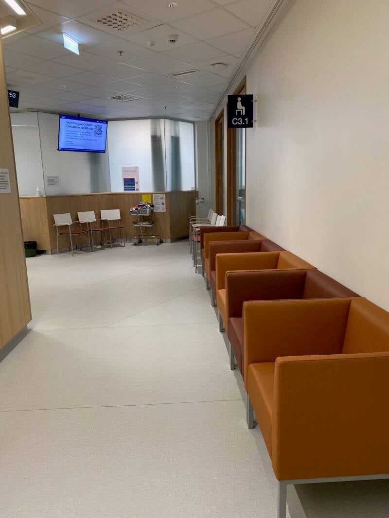 Syöpäkeskuksen lääkehoito-osaston odotushuone tyhjänä; vierekkäin monta tuolia ja vastapäisellä seinällä infoscreeni