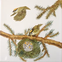 Ein handbemalter Porzellan-Pfannenuntersetzer zeigt ein Goldhähnchennest im Tannenbaum – die Mutter füttert, der Vater fliegt zur Futtersuche.