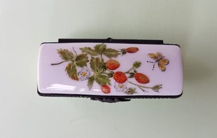 Diese längliche Pillendose zeigt auf dem Deckel eine Gruppe Erdbeeren mit Stiel, daneben fliegt ein Schmetterling – zart und lebendig bemalt.