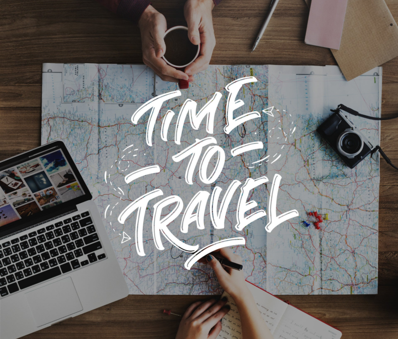 Time to Travel - Top view of a desk with maps, laptop, camera, notepad, and the hands of two people in front, organizing a trip