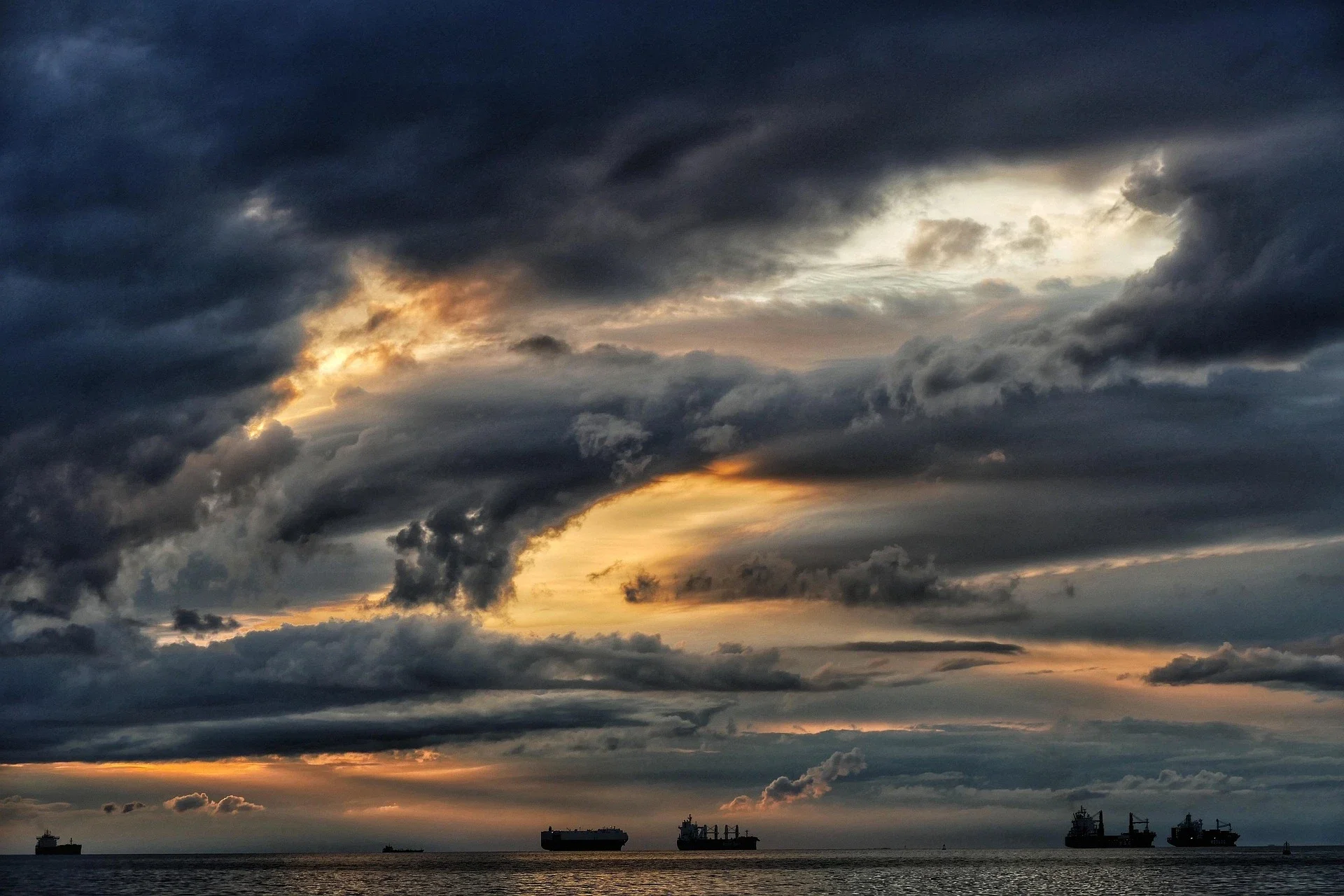 Donkere, grillige wolken hangen boven zee, terwijl gouden zonnestralen door openingen breken en vrachtschepen aan de horizon liggen.