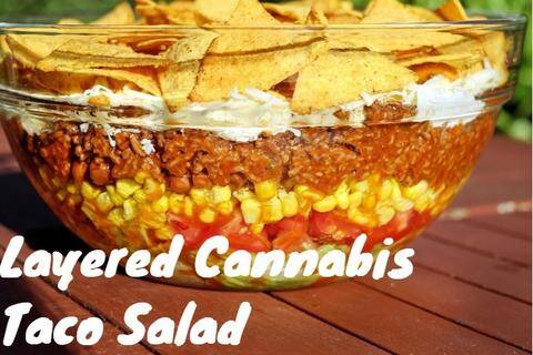 A beautiful layered cannabis taco salad in a glass bowl featuring lettuce, tomatoes, corn, beans, and seasoned ground beef topped with cheese and chips.