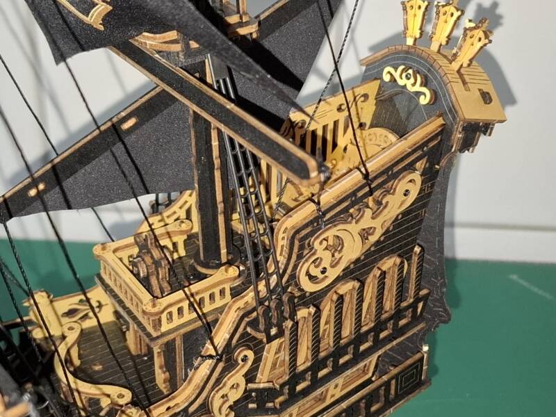 Gedetailleerd model van een piratenschip met zwarte zeilen en gouden schedel-emblemen, geplaatst op een houten sokkel. Het schip heeft meerdere masten, fijn tuigage en sierlijke ornamenten, en straalt vakmanschap en avontuur uit. Robotime MVB02