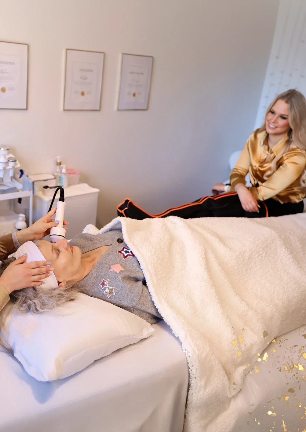 Kosmetologi tekee kasvohoitoa asiakkaalle hoitopedillä ja toinen asiakas rentoutuu lymfabootsit jaloissa hymyillen vieressä.