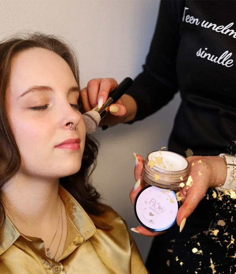 Meikkaaja levittää kevyttä päivämeikkiä asiakkaan kasvoille siveltimellä, korostaen luonnollista ja raikasta ilmettä.