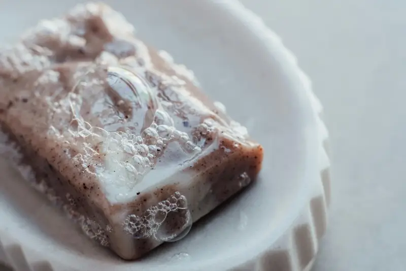 A Rectangle bar of exfoliating soap filled with fine, dark specks likely botanical seeds, clear soap bubbles and later coat the top and sides of the bar. The bar rests in a textured, off white ceramic dish, verticle ridges  and a grey background.
