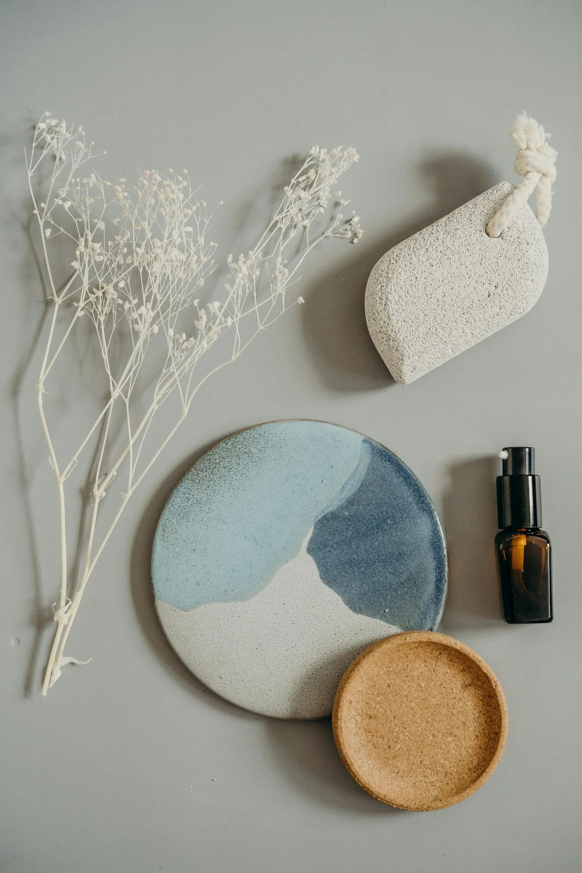 A flat lay composition on a solid grey muted surfact. An Asymmetrical Light grey volcanic rock with porous texture. A circular ceramic dish featuring an abstract glaze design, a small amber glass bottle,  A small bunch of white baby's breath 